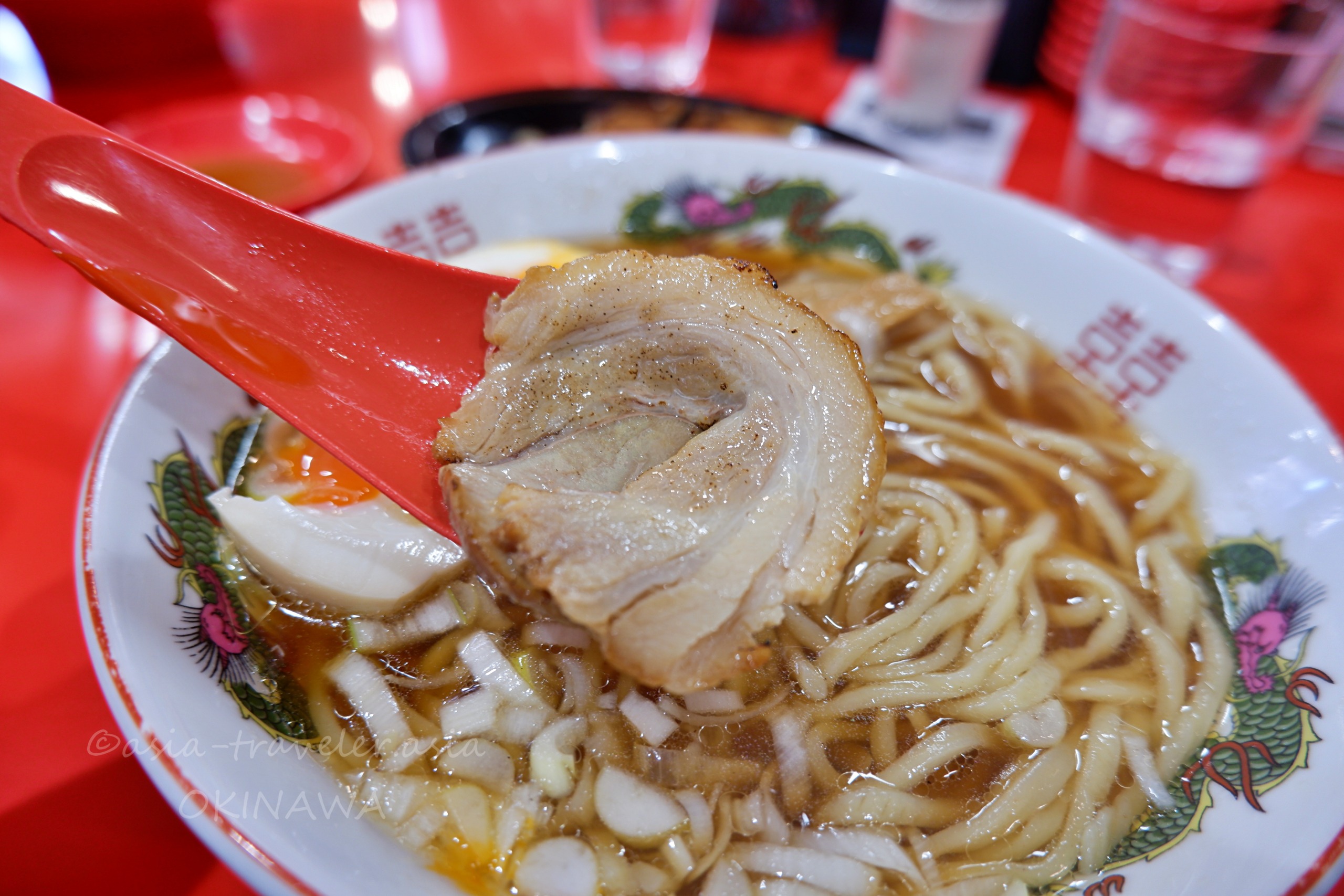 那覇Agu豚の赤いレンゲで持ち上げたチャーシューと味玉入りラーメン