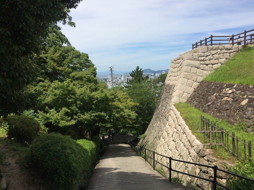 丸亀城の石垣沿いを下る参道と緑の木々・市街地の眺め
